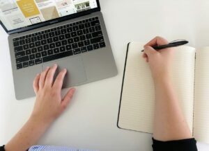 Hands writing in a notebook beside an open laptop